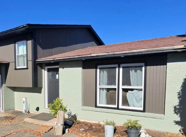 This is an image from A Plus Decorating Inc., located in Illinois. The image shows a residential home exterior with green painted brick, brown painted vertical siding, dark upper siding, white trim, and front windows.