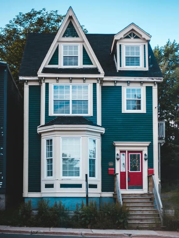 This is an image from Charlotte Pro Painters, located in North Carolina. This is a tall three-story home painted deep teal with cream trim and a red front door, highlighting exterior detail painting.