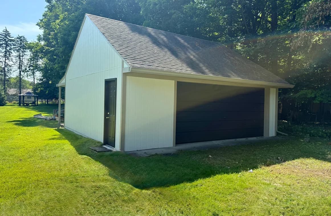 This is an image from MacFarland Painting - Ann Arbor company, located in Ann Arbor, MI The image is a freshly painted detached garage with white siding and dark brown garage door by MacFarland Painting, surrounded by green lawn and trees.