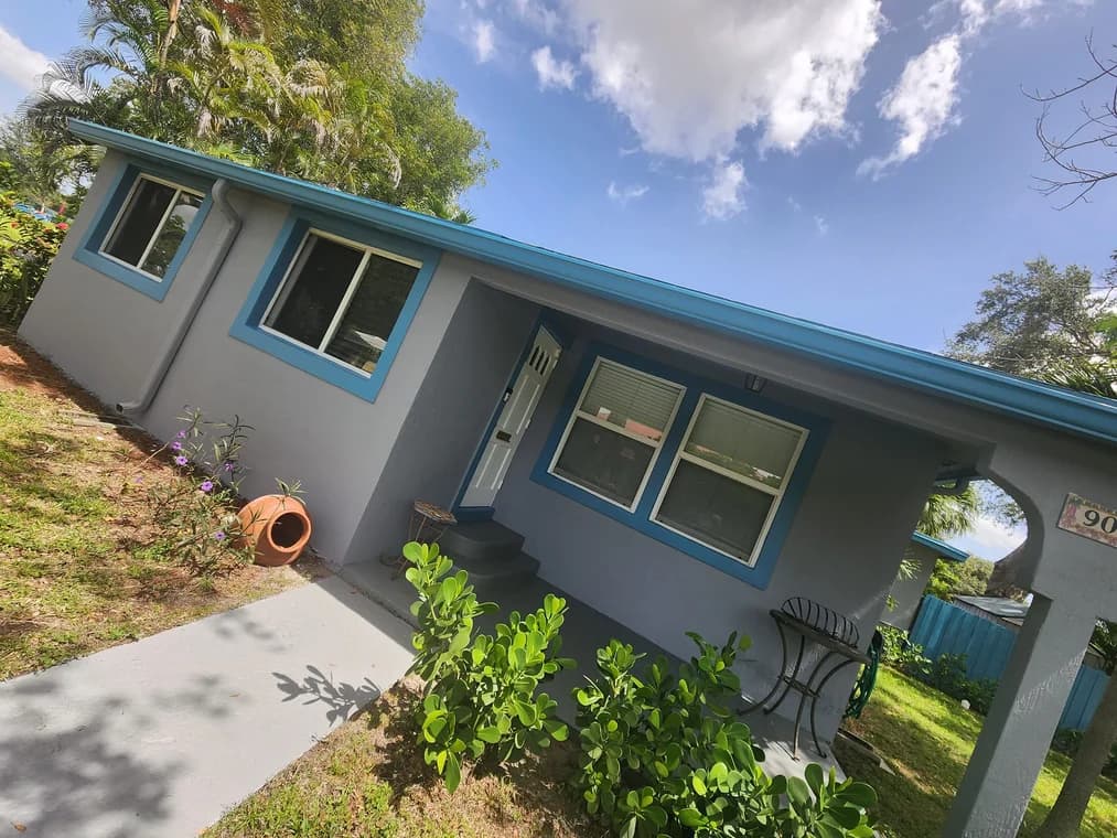This is an image from Big Family Painting Co  company, located in Boca Raton, FL. The image is a small single-story home exterior painted light gray with bright blue trim and roofline, featuring updated window frames and front porch
