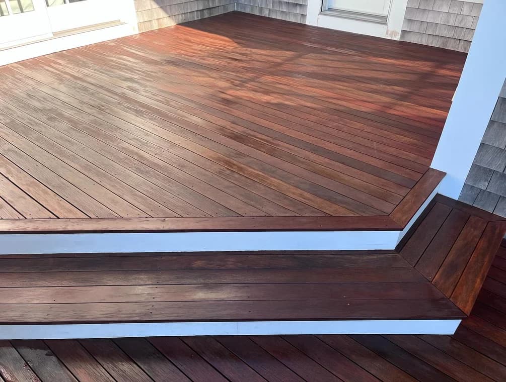 This is an image from Cape Cod Fine Finishes LLC, located in Massachusetts. It highlights a freshly refinished wooden deck with a rich brown stain, featuring clean lines and polished steps framed with white trim.