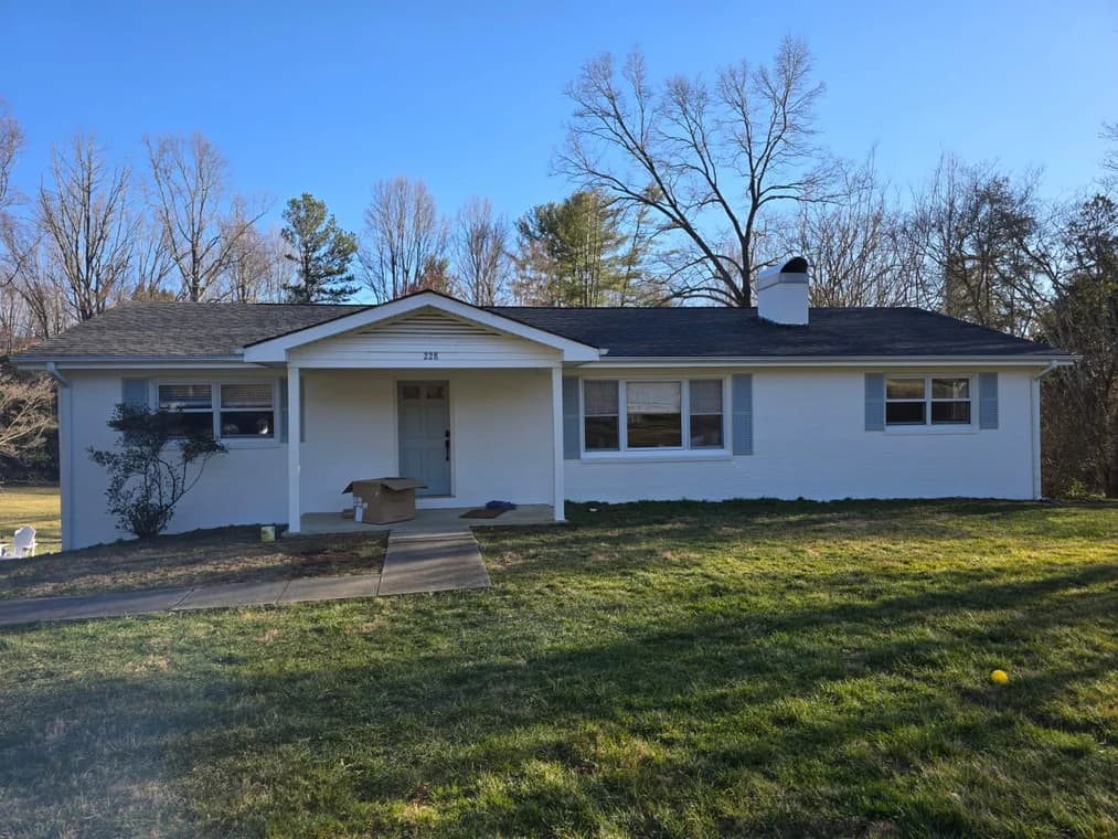 Exterior painting project by Custom Painting of Asheville inc in Asheville, NC. The image shows a white ranch home with blue shutters, front steps, lawn, and trees.
