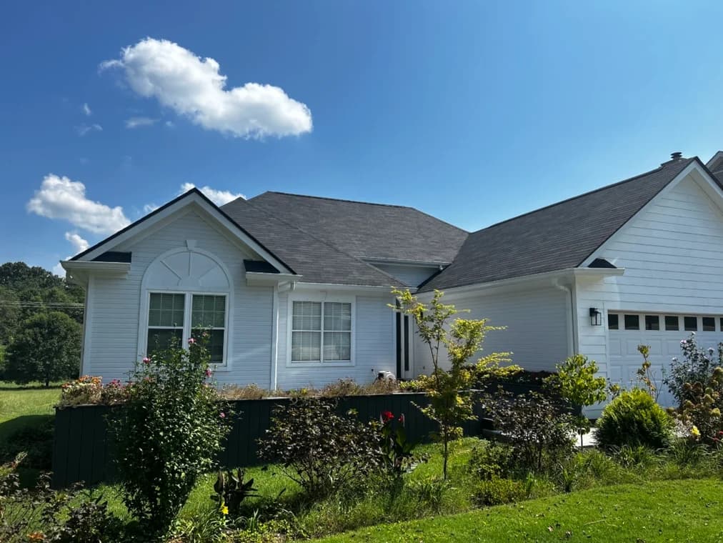 This is an image from Goodpaster Painting & Guttering, located in Tennessee. The image is a freshly painted white house with black roof and trim by Goodpaster Painting & Guttering under a clear blue sky.