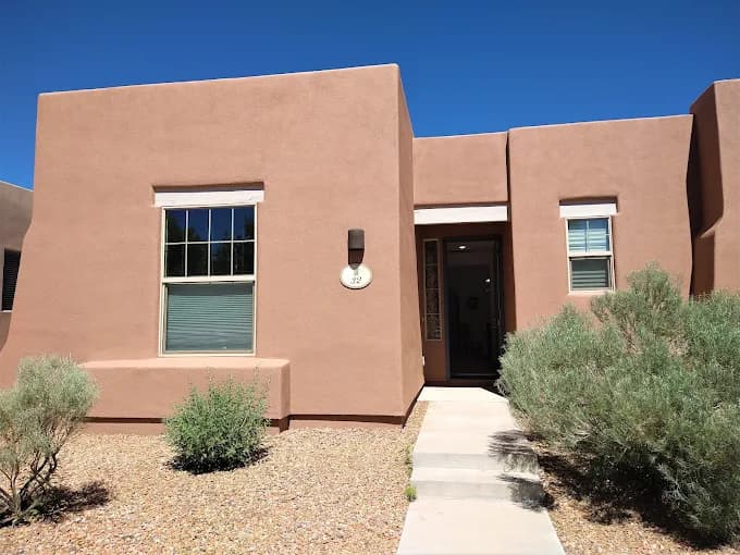 This is an image from Busby Finishes, located in New Mexico. The photo shows a modern stucco home with a smooth tan exterior finish, white trim, and desert landscaping under a clear blue sky.