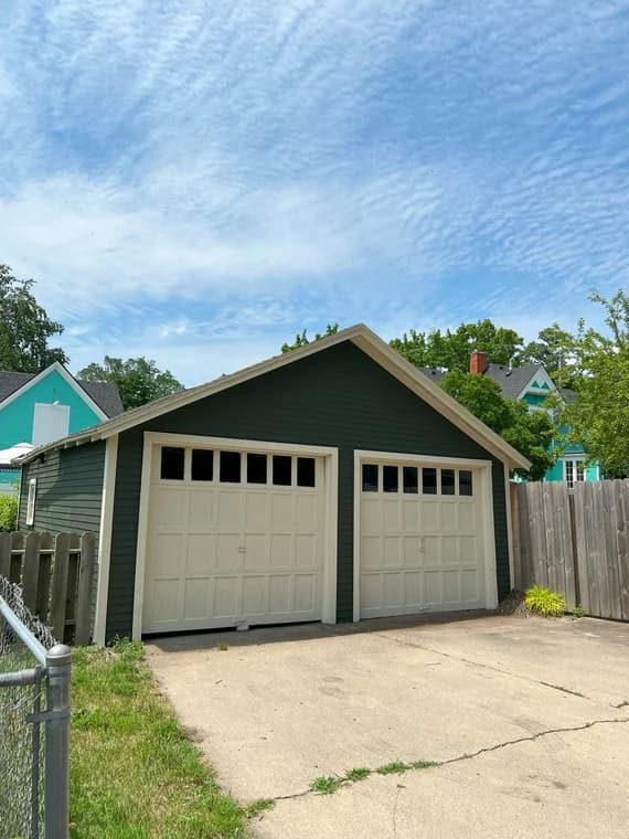 This is an image from Brushmen and Standby Bros company, located in Lansing, MI The image is a detached two-car garage with dark green siding and cream trim, freshly painted by Brushmen and Standby Bros, residential exterior painting project.
