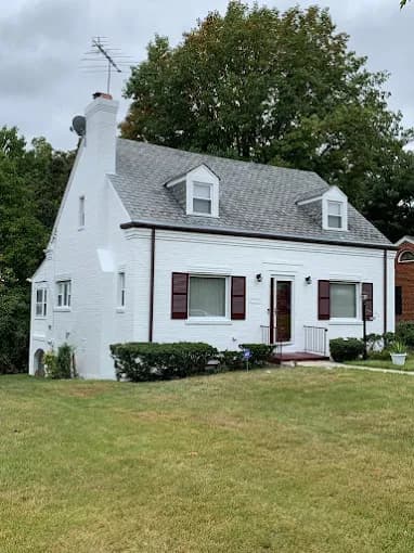 This is an image from CertaPro Painters of Southern Maryland, located in Maryland. The photo shows a freshly painted white brick home with dark red shutters, a gray roof, and a neatly trimmed front yard.