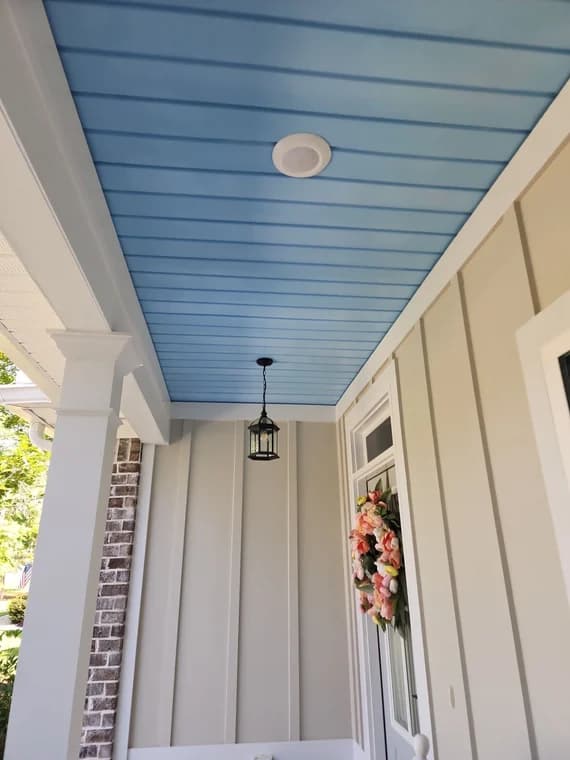 This is an image from A Cut Above Painting Co. company, located in Evans, GA The image is a front porch with light blue beadboard ceiling, beige board and batten siding, and floral wreath on white door