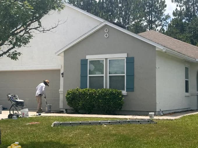 This is an image from Rockstar Painting, Inc. company, located inJacksonville, FL. The image is an exterior house painting in progress by professional painter—single-story home with gray and white facade, teal window shutters, and painting equipment on lawn.