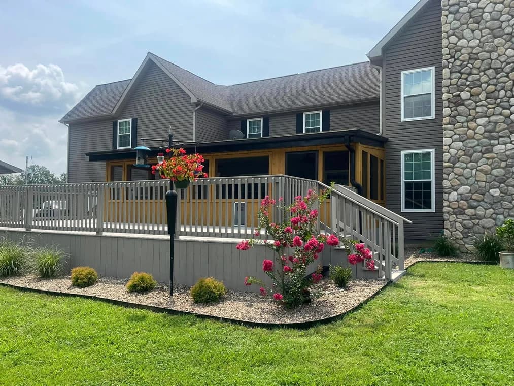 This is an image from Rodgers Painting, located in Kentucky. The exterior of a large two-story home with taupe siding, a stone chimney, and a light gray deck surrounded by a landscaped garden is shown under a bright sky.