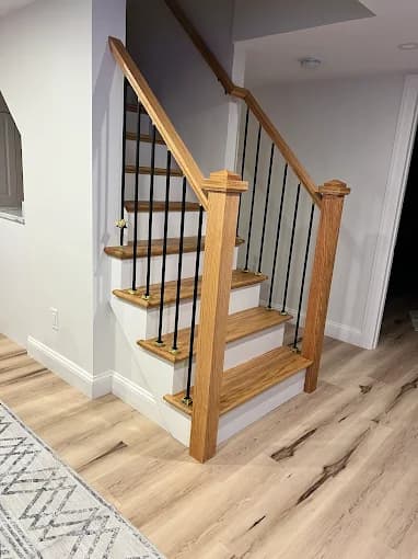 This is an image from Elite Painting Services Plus, located in New York. The photo shows an indoor staircase with wooden steps, black metal balusters, and oak newel posts in a newly finished interior space.