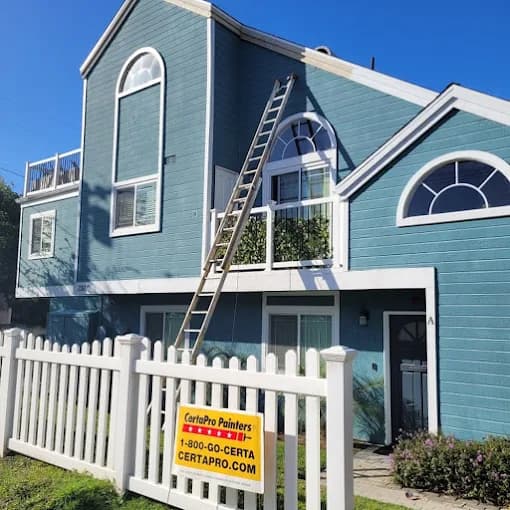 This is an image from CertaPro Painters of Attleboro, MA. It shows a two-story blue house with white trim, a white picket fence, and a tall ladder placed against the exterior during painting.