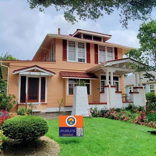 This is an image from Pelican Paint Group, located in Louisiana. The photo shows a freshly painted two-story home with peach siding, white trim, and red shutters, surrounded by a well-kept lawn and flowers with a Pelican Paint Group yard sign displayed in front.