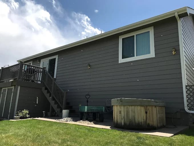 This is an image from Noble Painting company, located in Fort Collins, CO The image is a rear exterior of a single-story home with gray horizontal siding, white trim, elevated wood deck with stairs, hot tub, and freshly painted finish by Noble Painting