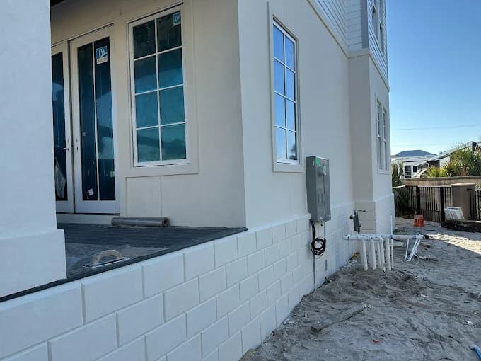 This is an image from Falcon Remodeling Express LLC company, located in Fort Walton Beach, FL. The image is a newly painted stucco exterior of a modern coastal home with raised foundation, double French doors, and clean white finish near sandy terrain