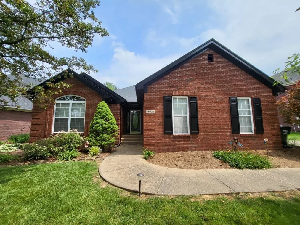 This is an image from Gomez Painting LLC, located in Kentucky. The photo shows a red brick house with black shutters, a manicured front yard, and a curved concrete walkway leading to the entrance.