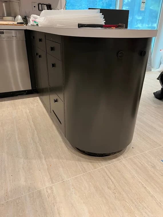 This is an image from Nyca Contractors, LLC, located in New York. The photo shows a dark modern kitchen island with curved cabinetry, matte black panels, and a light countertop installed over large beige floor tiles.