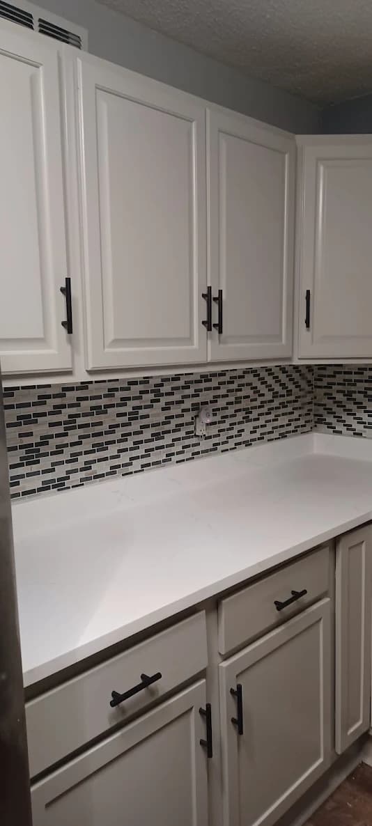This is an image from Scarlett Painting & Remodeling. The image shows freshly painted white kitchen cabinets with a clean, updated finish and a modern mosaic tile backsplash.