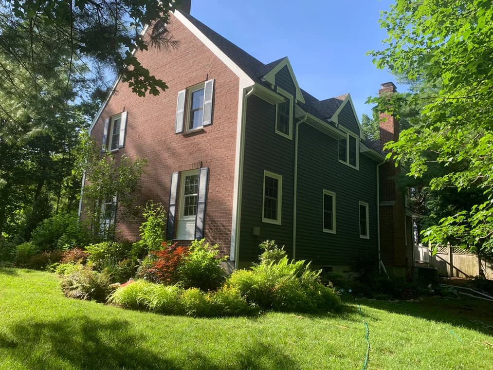 This is an image from Decolores Painting, featuring a traditional brick house with dark green painted siding and blue shutters. The home is surrounded by a landscaped garden and trees.
