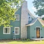 This is an image from West & Co Painting, LLC. The image shows a blue-green house with white trim and a tall stone chimney surrounded by trees.