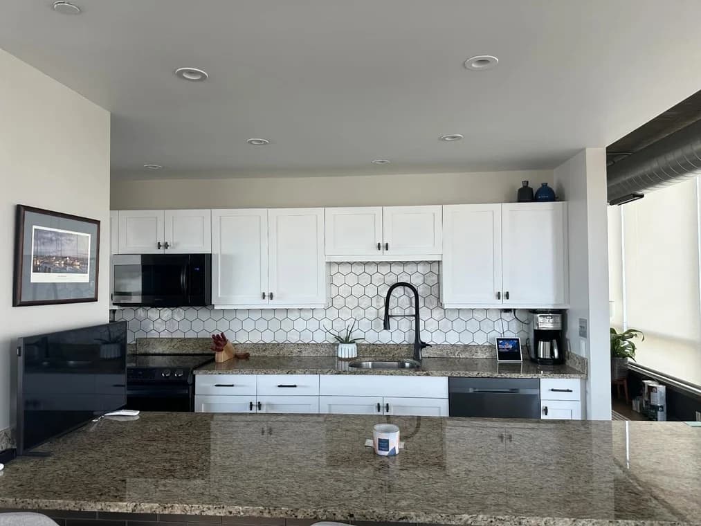This is an image from Paint Perfection, located in Maryland. The picture shows a bright kitchen with white cabinets, hexagon tile backsplash, granite countertops, and stainless appliances.