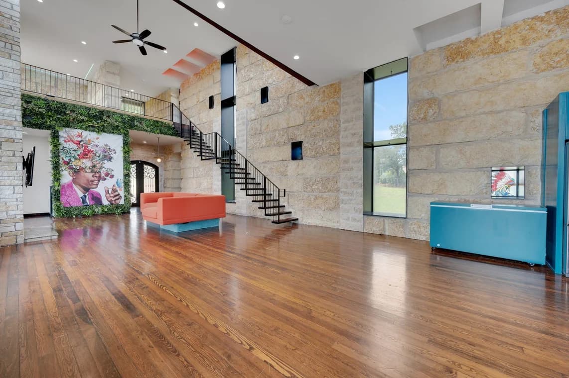 "This is an image from Reyes Painting Co., located in Texas. The interior image displays a large open living space with stone walls, tall windows, hardwood floors, a staircase, and a colorful art feature framed by greenery."