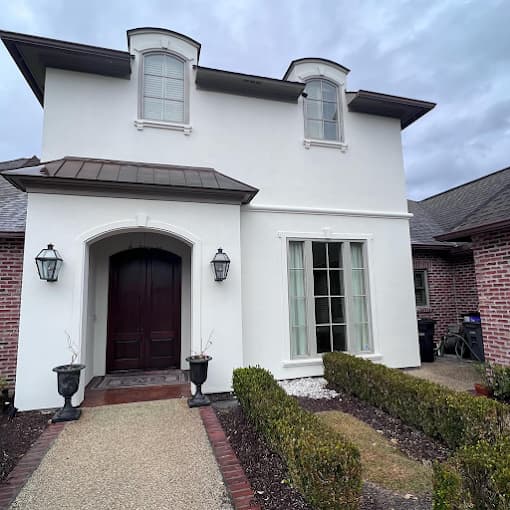 This is an image from Yeras Painting, located in Louisiana. The photo shows a freshly painted white stucco home with dark wood doors, light trim, and neatly trimmed hedges leading up to the entrance.