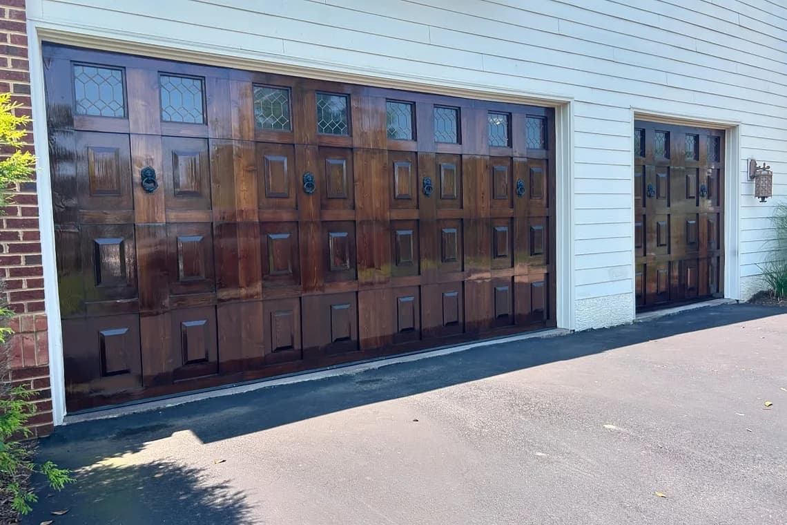 This is an image from MK Painting & Custom Finishes Co company, located in Schwenksville, PA. The image is a refinished wood garage doors with rich stain, raised panel design, and upper leaded glass windows by MK Painting