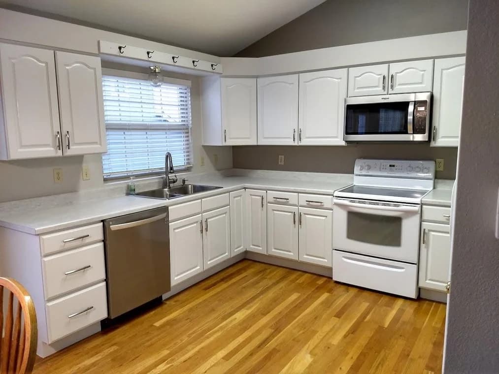This is an image from All Painting Plus 2, LLC company, located in Colorado Springs, CO The image is a modern kitchen with freshly painted white cabinets, stainless steel appliances, wood flooring, and bright natural lighting – All Painting Plus 2 LLC cabinet painting project.