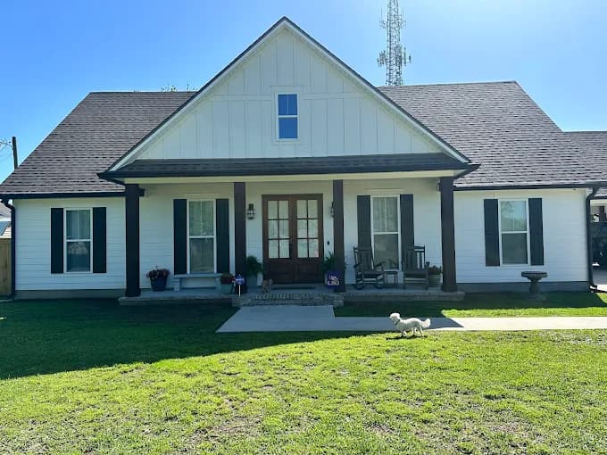 This is an image from Yeras Painting, located in Louisiana. The photo shows a single-story white farmhouse-style home with dark brown trim, a covered front porch, and a small dog walking across the green lawn in front.