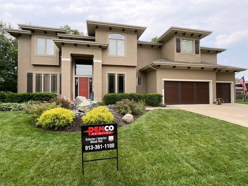 This is an image from PEMCO Painting Interior & Exterior Painter Overland Park. The image is PEMCO-Painting-Interior-&-Exterior-Painter-Overland-Park-1.webp exterior house in Kansas City KS with freshly painted stucco walls and dark wood-tone garage doors.