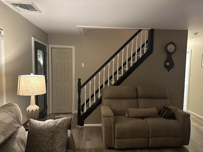 This is an image from Big Expert Painting LLC, located in New York. The interior photo shows a living room with freshly painted beige walls, a staircase with black trim and white spindles, and updated modern lighting.