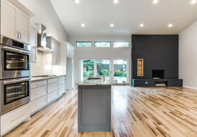 This is an image from Big Al's Painting, located in Texas. The photo shows a spacious open-concept kitchen and living area with light wood flooring, white cabinetry, stainless steel appliances, and a dark accent fireplace wall.