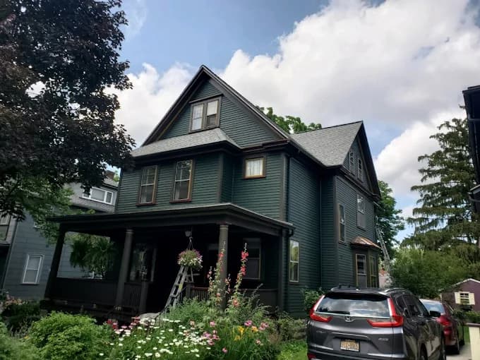 This is an image from Monarch Painting, located in New York. The photo shows a newly painted dark-green two-story home with detailed trim work, a covered porch, and garden landscaping in the front yard.