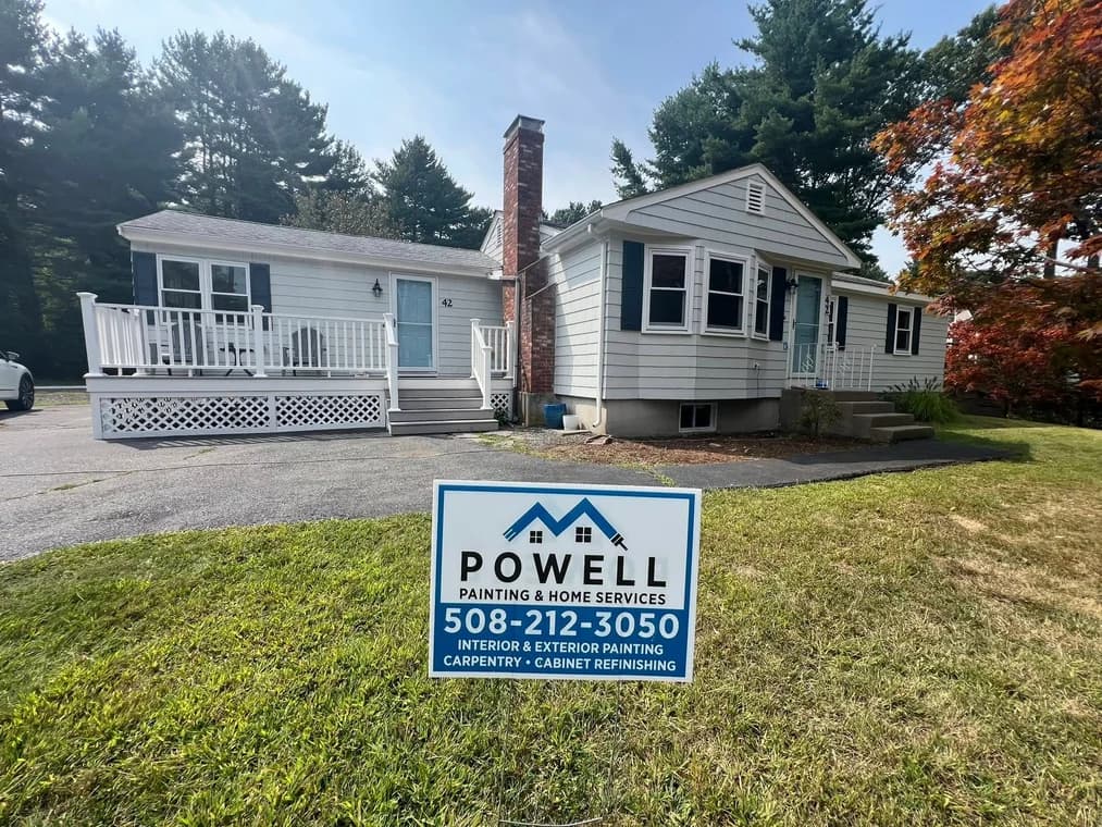 This is an image from Powell Painting & Home Services, located in Massachusetts. The photo shows a freshly painted light gray home with white trim, a blue front door, and a Powell Painting yard sign displayed in the front lawn