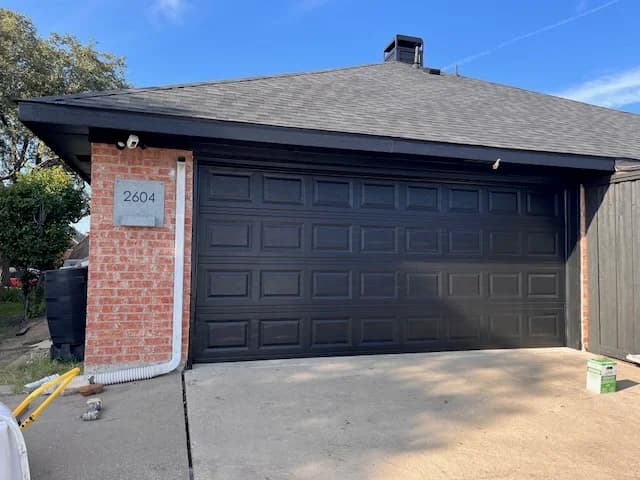 This is an image from Platinum Painting of Richardson, located in Texas. The driveway view shows a freshly painted black garage door on a brick home with the house number 2604 displayed on a mounted plaque.