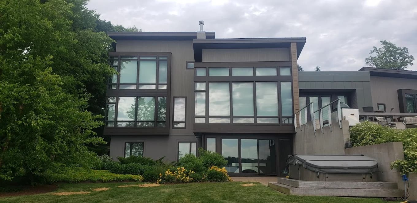 This is an image from Braun's Painting, LLC, located in New York. The photo shows a modern multi-story home exterior with dark trim, expansive windows, and freshly painted siding surrounded by greenery.