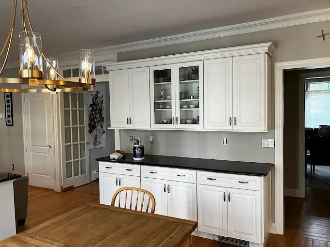 This is an image from Anthony's Painting & Decorating, Inc., located in New York. The photo shows a dining area with freshly painted white cabinetry, glass-front upper doors, and a dark countertop against light gray walls.