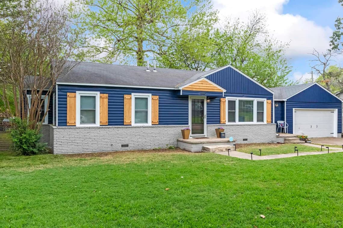 This is an image from Impressions Painting, located in Tulsa, Oklahoma. The photo shows a single-story home with navy blue siding, light gray painted brick, natural wood shutters, and a white garage door.