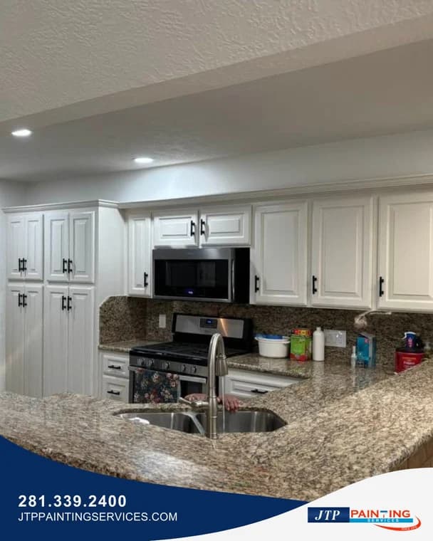 This is an image from JTP Painting Services, located in Texas. The photo shows a bright kitchen with white painted cabinets, stainless steel appliances, and granite countertops.