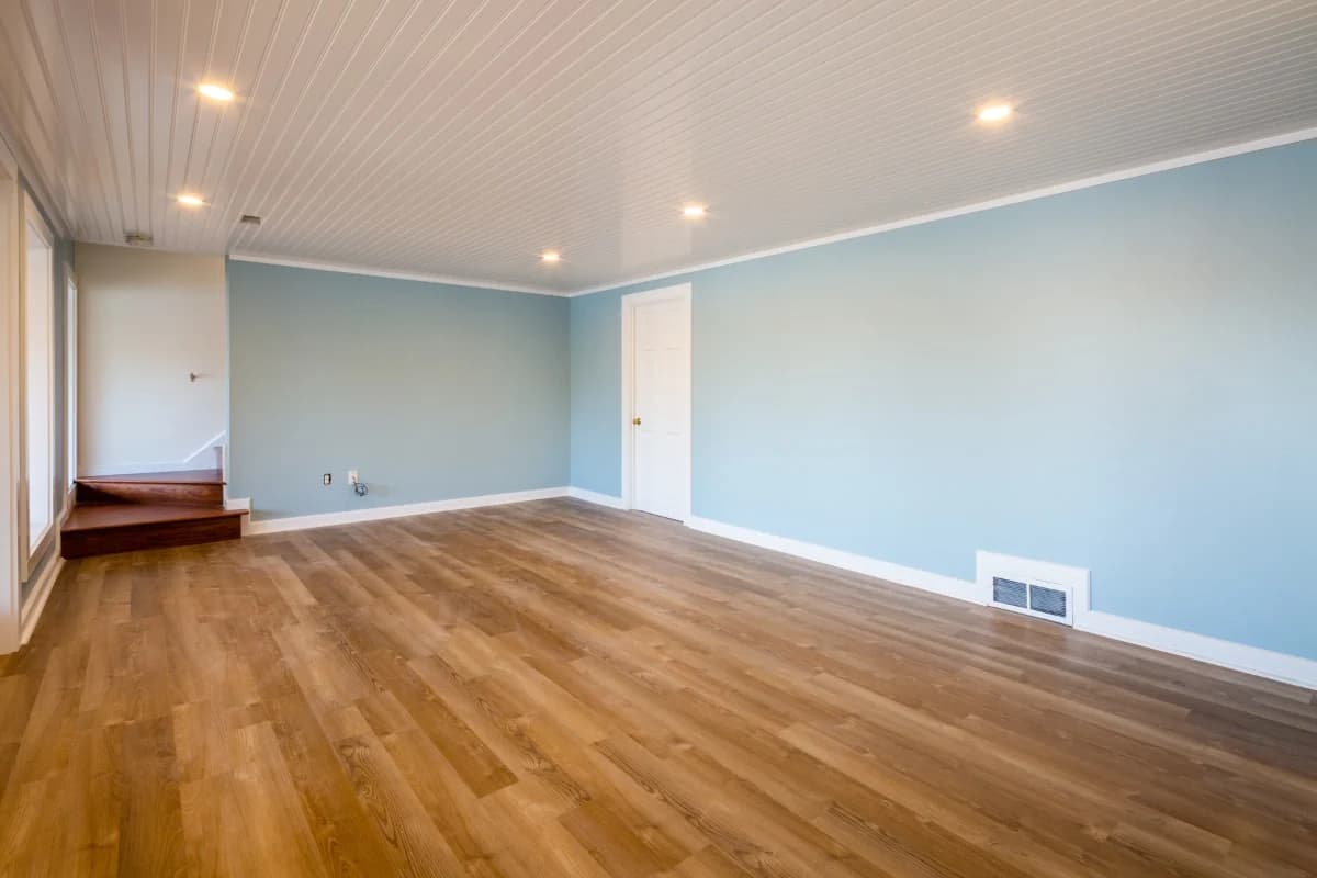 This is an image from River City Painting Inc. The image shows a bright basement room with blue painted walls, white trim, and wood-look flooring.