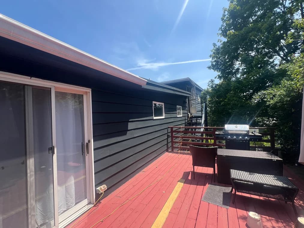 This is an exterior project by Rodrigo's Painting. The house siding is painted black, contrasted with a red deck and wooden railing, with patio furniture and a barbecue on the deck.