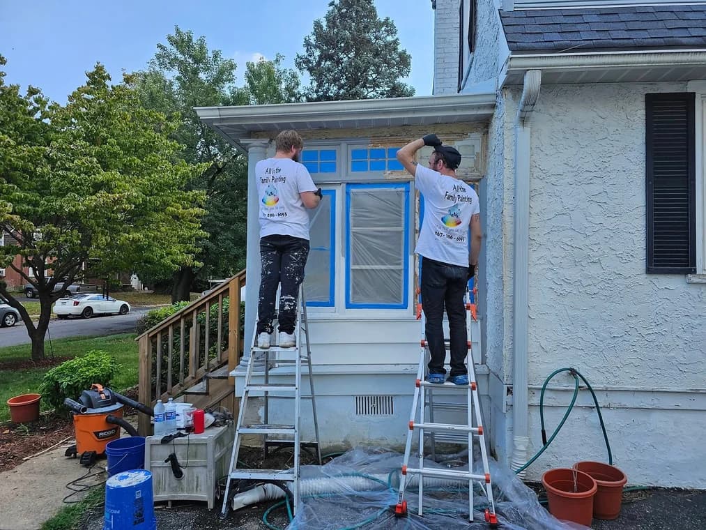 This is an image from All in the Family Painting company, located in Brookhaven, PA. The image is a two painters from All in the Family Painting preparing a house exterior for painting, wearing branded shirts and working on ladders with taped windows and protective coverings.
