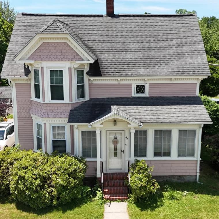 This is an image from Paint Bros MA, located in Massachusetts. It features a two-story Victorian-style home painted in light pink with white trim, showcasing its detailed windows, gable roof, and welcoming front entry.