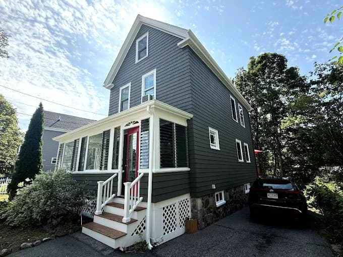 This is an image from JK Painting Service, located in Massachusetts. The photo shows a freshly painted dark gray two-story home with white trim, a red front door, and a stone foundation, highlighting the company’s exterior painting work.