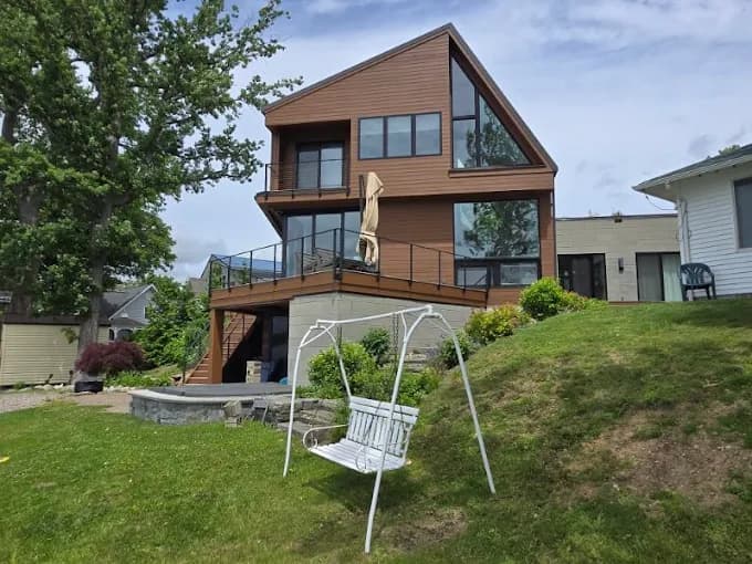 This is an image from Level Up Painting, located in New York. The photo shows a modern multi-level lakefront home with brown exterior siding, large glass windows, and black trim overlooking a grassy yard.