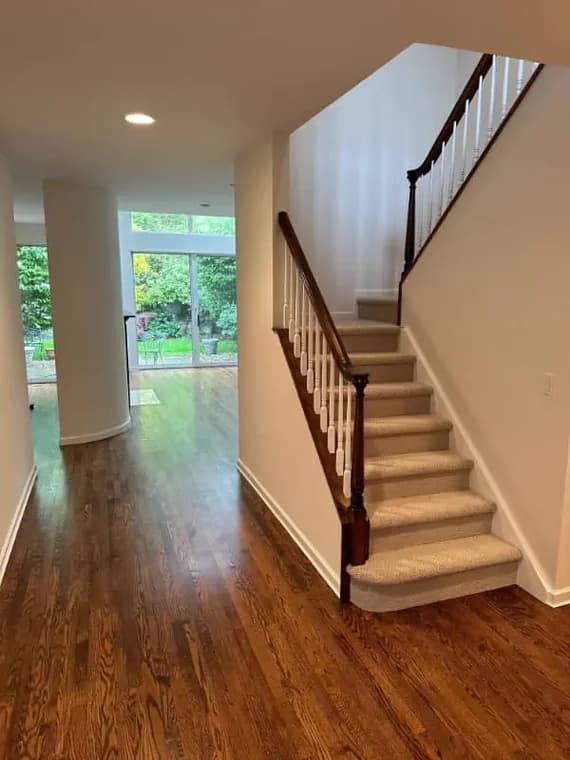 This is an image from Pier Painting Services, located in NY. The hallway features newly painted neutral walls, refinished hardwood floors, and a staircase with white risers and carpeted steps.