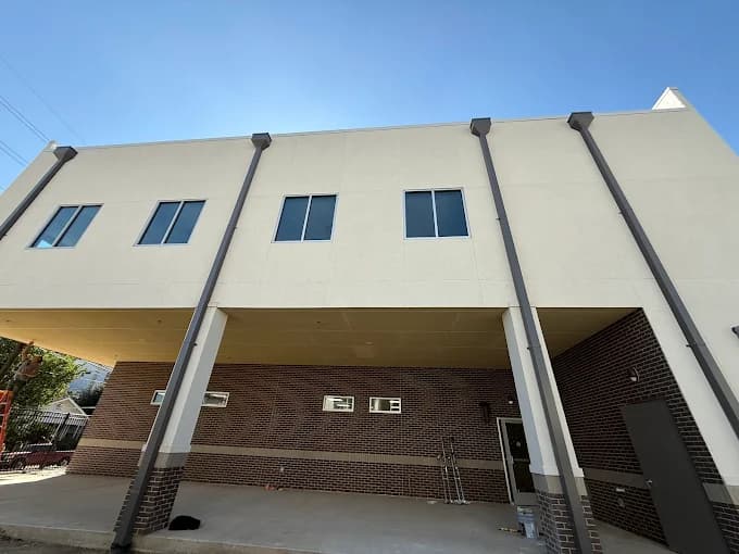 This is an image from Blue Painting, located in Texas. The image shows a commercial building exterior with light-colored upper walls, dark brick lower walls, tall vertical support columns, and a covered drive-through area under a clear blue sky.