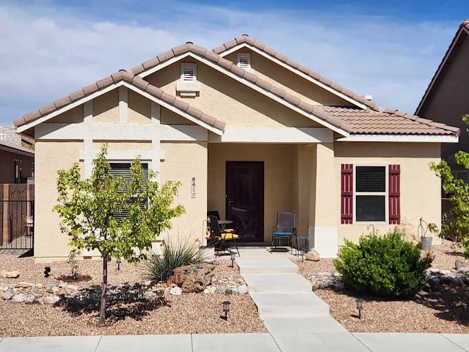 This is an image from Mike’s Pro Painting, LLC, located in New Mexico. The photo shows a freshly painted tan stucco home with a tile roof, white trim accents, red shutters, and desert landscaping under a clear blue sky.