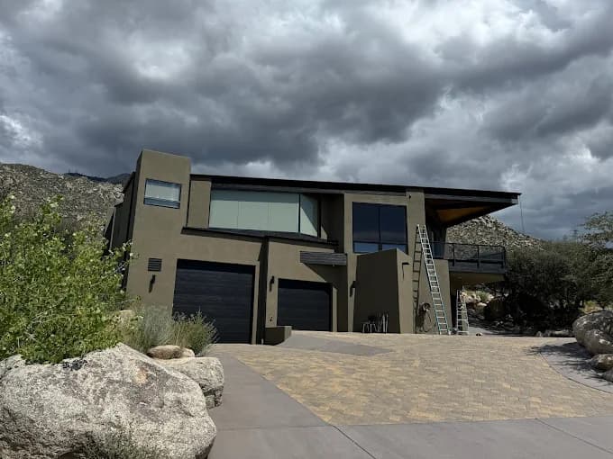This is an image from Straight Line Painting LLC, located in New Mexico. The photo shows a modern two-story home with a fresh dark exterior finish under dramatic cloudy skies, with ladders and painting equipment visible on the driveway.