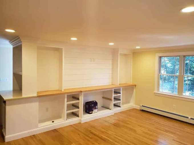 This is an image from WM Painting & More, located in Massachusetts. The photo shows a freshly painted living space with cream-colored walls, built-in shelving, recessed lighting, and hardwood floors.